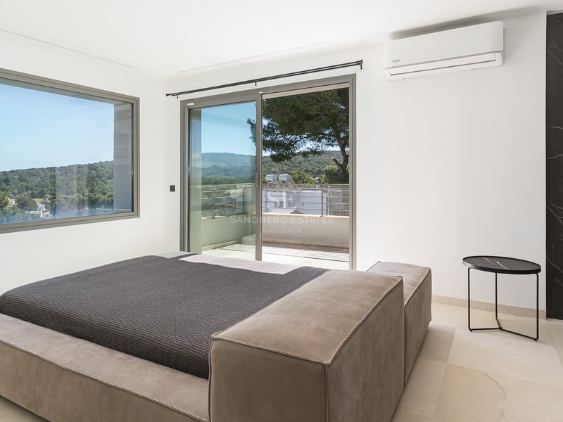 Modernt sovrum med låg sängram, stora skjutdörrar i glas mot terrass och fönster med utsikt över gröna kullar.