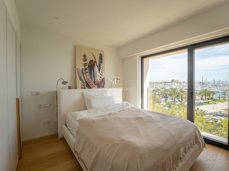 Modernt sovrum med vit säng och ett stort fönster med utsikt över en solig marina med palmer.