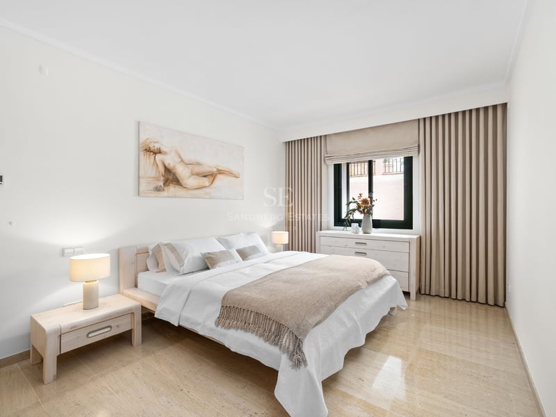 Chambre lumineuse avec lit en bois clair, sol en marbre, murs blancs et un grand tableau au-dessus du lit.