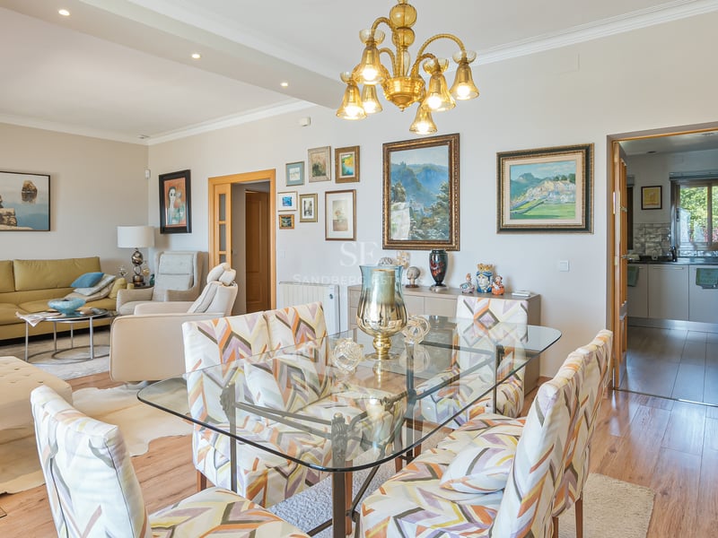 Ljust rum med öppen planlösning, matbord i glas, mönstrade stolar och konstsamling på väggen.