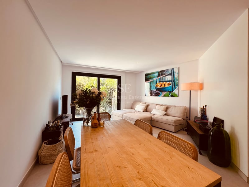Salon et salle à manger avec grande table en bois, chaises en rotin et canapé beige près d'une baie vitrée.