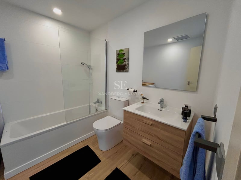 Contemporary bathroom with white walls, glass-screened bathtub, toilet, and light wood vanity unit.