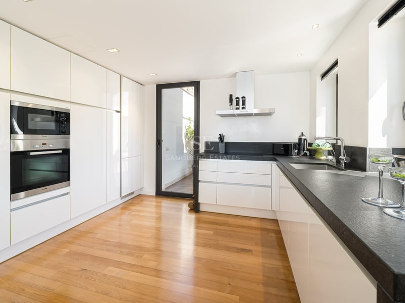 Modern white kitchen with dark stone countertops, oak flooring, integrated appliances, and access to a terrace.