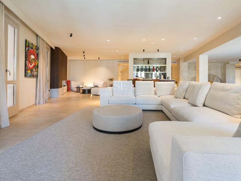 Espace de vie moderne et spacieux avec un grand canapé d'angle blanc, un tapis neutre et un bar élégant au fond.