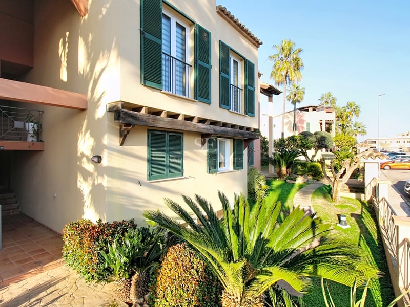 Exteriör av en beige byggnad med gröna fönsterluckor, träbjälkar och tropisk trädgård.