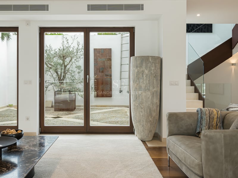 Salon moderne avec canapé en cuir, table en marbre et grandes baies vitrées ouvrant sur un patio avec un olivier.