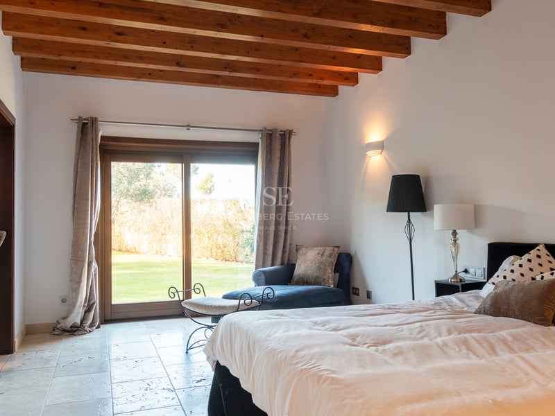 Sovrum med synliga takbjälkar i trä, naturstengolv och skjutdörr i glas mot trädgården.