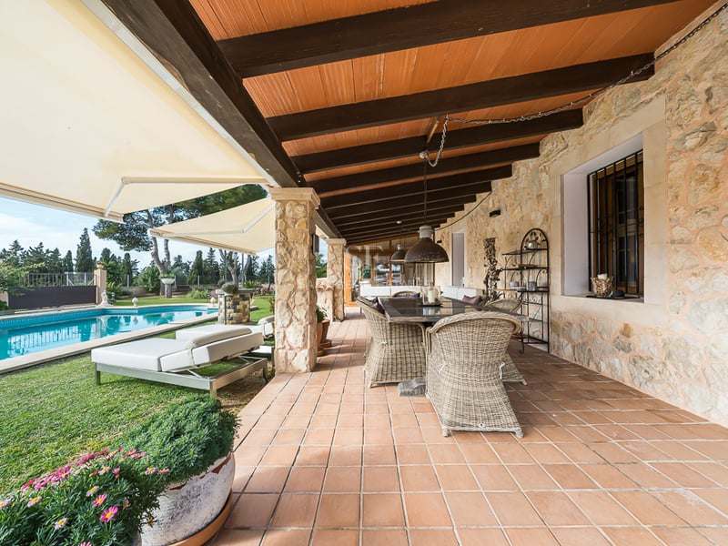 Grande terrasse couverte avec carrelage en terre cuite, murs en pierre et salle à manger en osier face à la piscine.