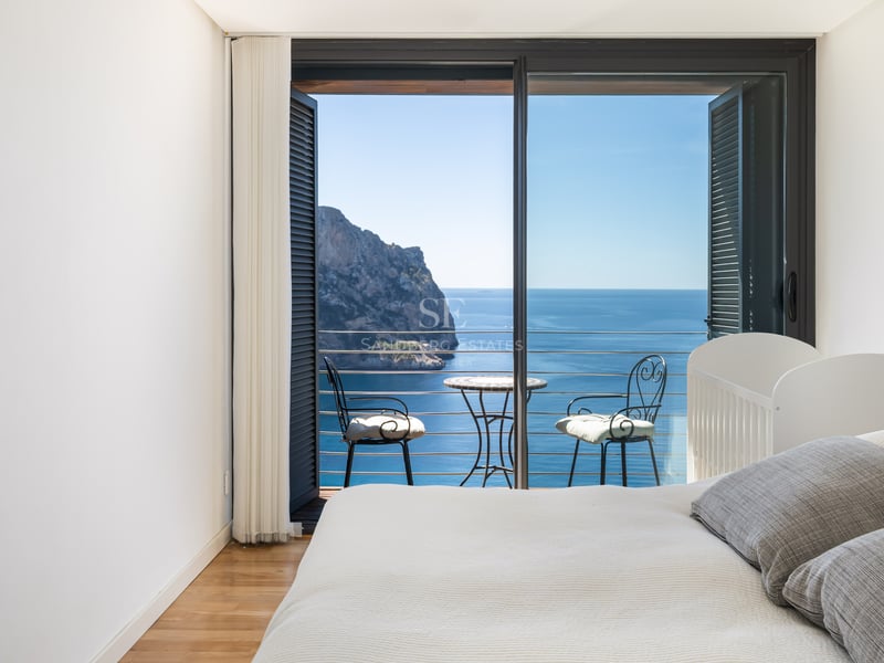 Minimalist white bedroom featuring a private balcony with wrought iron furniture and front-on views of a sea cliff.