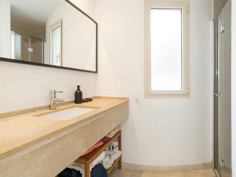 Modernt badrum med ett långt stentvättställ i beige, stor spegel och duschvägg i glas i ett ljust rum.