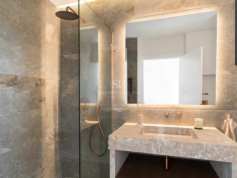 Luxuriöses Badezimmer mit begehbarer Glasdusche, Steinwaschtisch und großem hinterleuchtetem Spiegel mit warmem Licht.