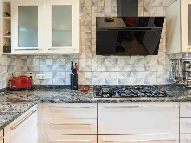 Närbild av ett modernt kök med bänkskiva i granit, gasspis, mönstrat kakel och vita skåpluckor.