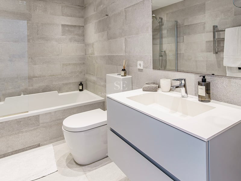 Contemporary bathroom featuring grey stone-effect tiles, a white bathtub, toilet, and modern vanity with a large mirror.