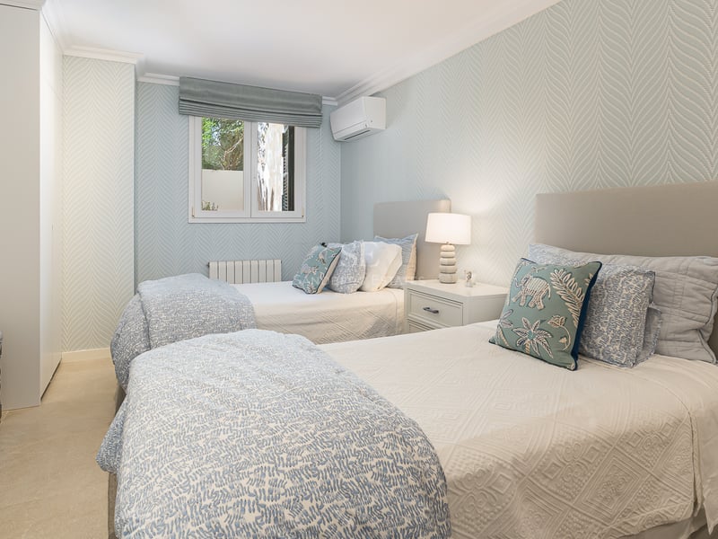 Bright room with two single beds, light blue patterned wallpaper, built-in wardrobes, and stone flooring.