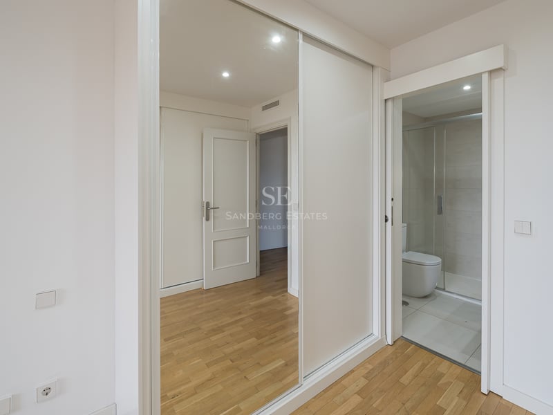 Vy av ett modernt sovrum som visar skjutdörrar med speglar till en garderob och en öppning till ett vitt modernt badrum.