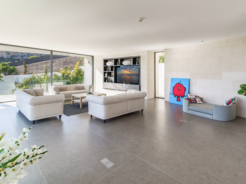 Salon moderne et lumineux avec canapés beiges, sol en pierre grise et grandes baies vitrées donnant sur un jardin.