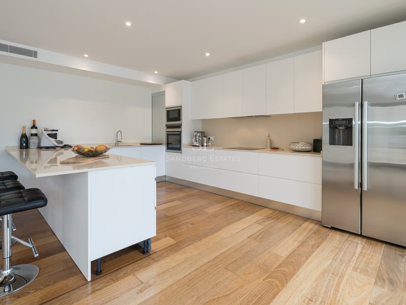 Cuisine blanche moderne avec parquet, réfrigérateur en inox, plan de travail clair et tabourets de bar noirs.