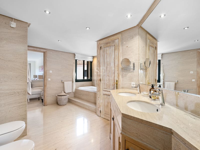 Salle de bain spacieuse avec murs en pierre beige, double vasque, grand miroir et baignoire encastrée au fond.