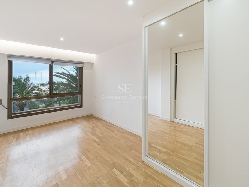 Tomt sovrum med ljust trägolv, spegelgarderob och fönster med utsikt över palmer och båtar.