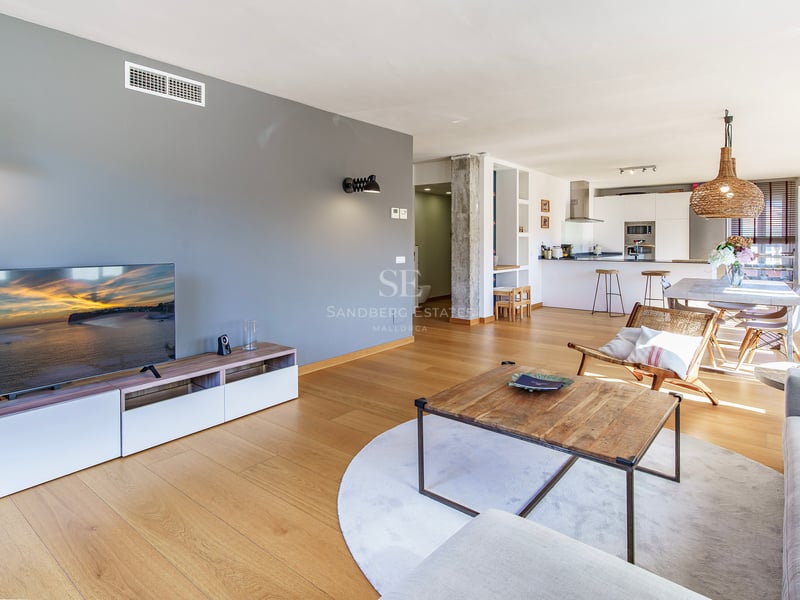 Ljust vardagsrum med öppen planlösning, ljusa trägolv, grå fondvägg och integrerat kök i bakgrunden.