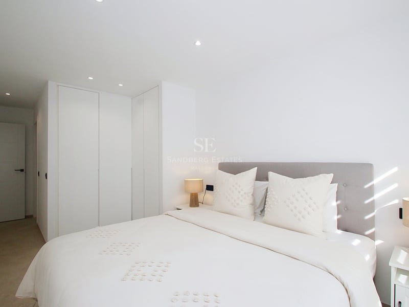 Ett ljust modernt sovrum med vita sängkläder, grå huvudgavel, integrerade garderober och naturligt ljus.