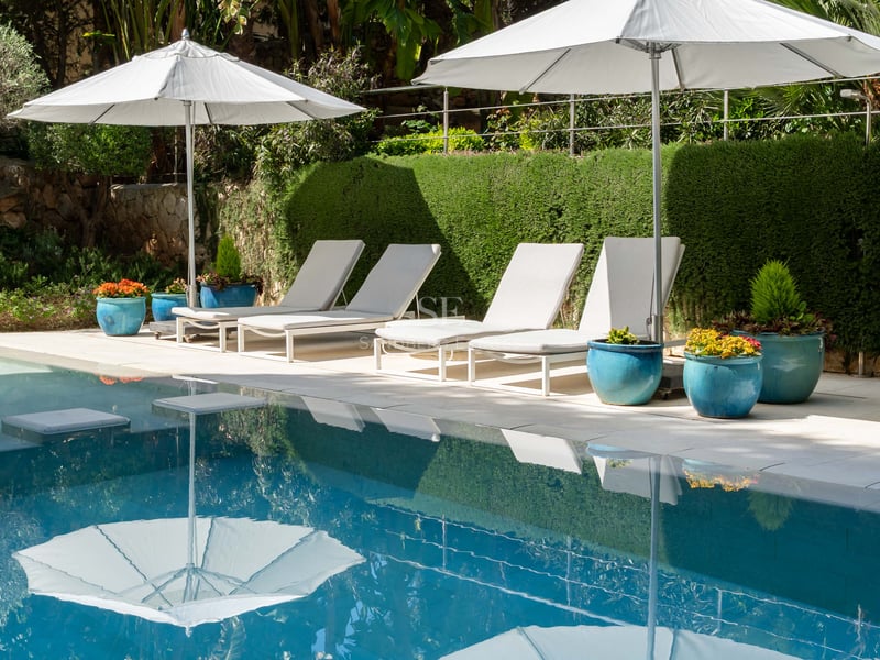 Luxuriöser Swimmingpool mit vier weißen Liegestühlen und Sonnenschirmen vor einer gepflegten grünen Hecke.