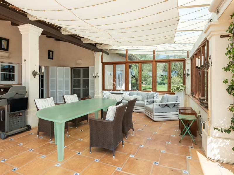 Terrasse couverte avec sol en terre cuite, table à manger, coin salon et barbecue sous un auvent en toile.