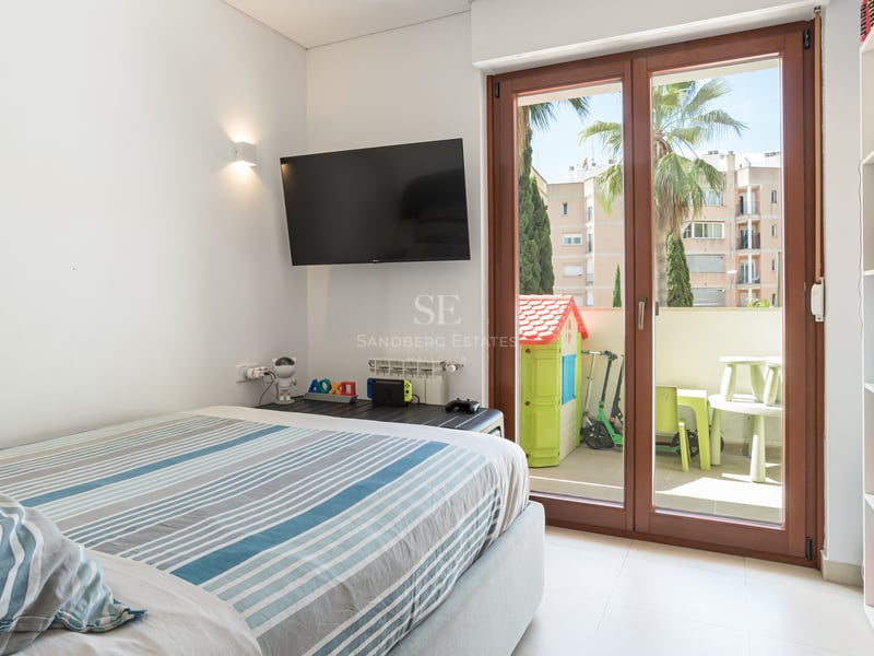 Chambre lumineuse avec lit à rayures, TV murale et baies vitrées ouvrant sur une terrasse avec jeux d'enfants.