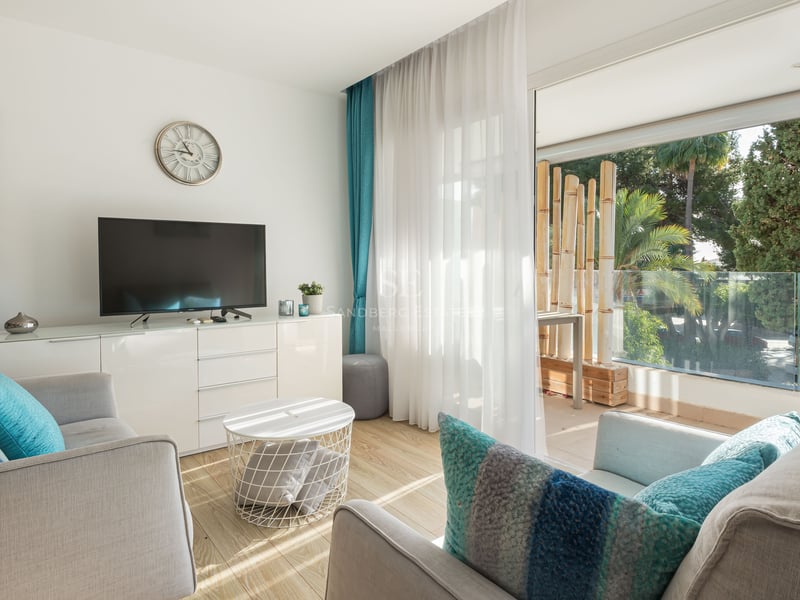 Salon moderne avec canapés gris, coussins turquoise et grandes baies vitrées ouvrant sur une terrasse arborée.