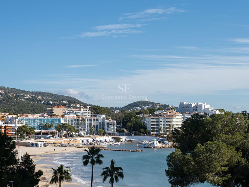 Vue dégagée sur une ville côtière avec des bâtiments blancs, une plage de sable, des palmiers et un petit port sous un ciel bleu.