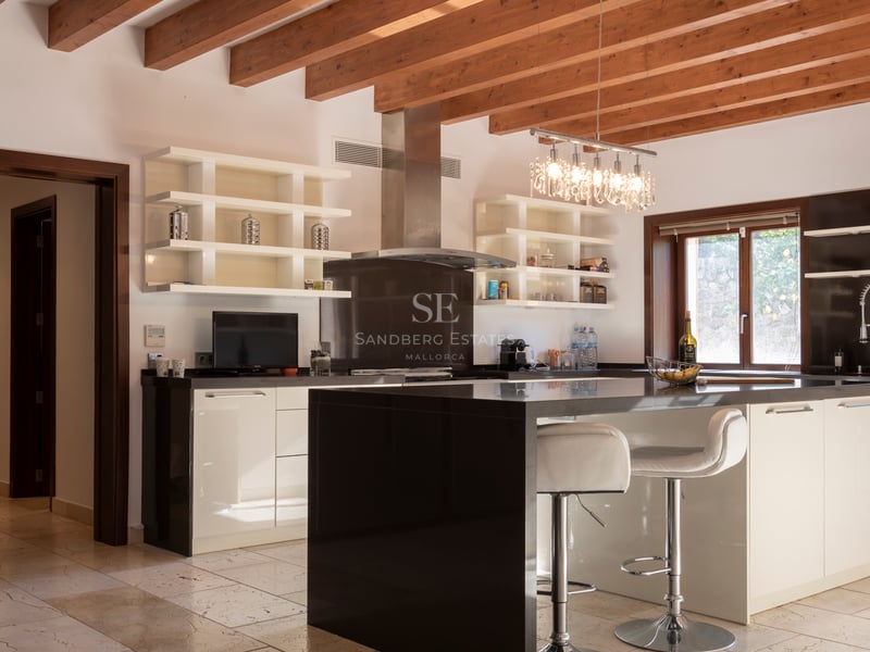 Modernt kök med svart köksö, vita skåp, synliga träbjälkar och naturstensgolv.