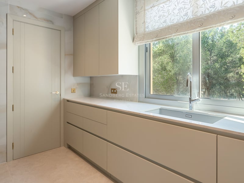 Minimalist kitchen featuring cream handleless cabinets, a large window overlooking greenery, and a stone backsplash.