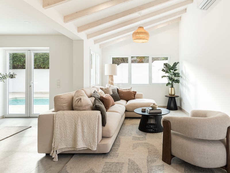 Salon lumineux avec canapé beige, plafond à poutres de bois et portes vitrées ouvrant sur une piscine.