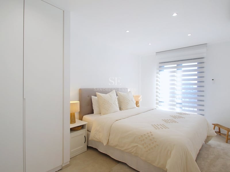 Välbelyst modernt sovrum i neutrala färger med dubbelsäng och vita inbyggda garderober.