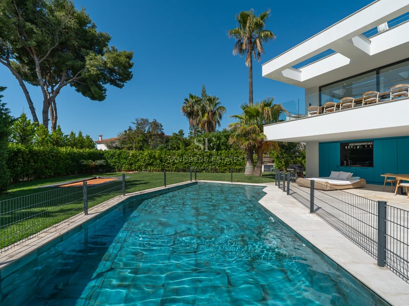 Villa blanche moderne avec piscine bleue, meubles de terrasse en bois et jardin paysager sous un ciel bleu.
