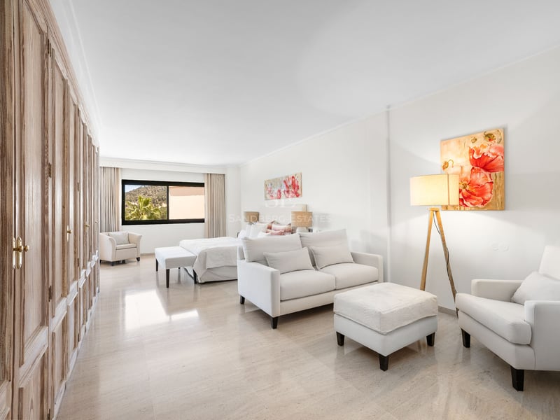 Suite lumineuse avec canapé blanc, armoires en bois, grand lit et art floral sur sol en marbre poli.