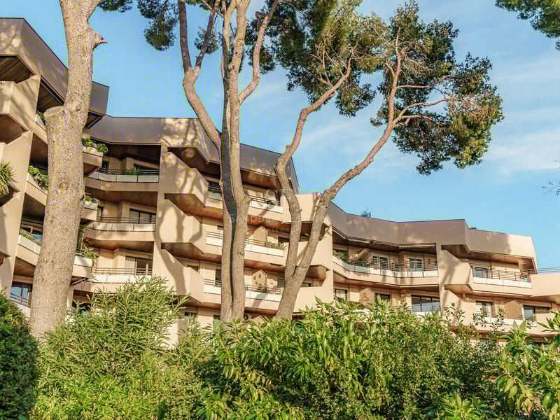Mehrstöckiges modernes Gebäude mit eckigen Balkonen, umgeben von hohen Pinien unter klarem blauem Himmel.