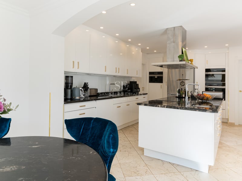 Zeitgenössische weiße Küche mit schwarzen Steinarbeitsplatten, Kochinsel und blauem Samtstuhl.