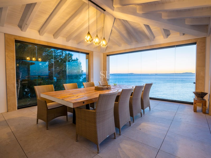 Großer Holzesstisch auf einer Terrasse mit Glasvorhängen und Blick auf das blaue Meer bei Dämmerung.