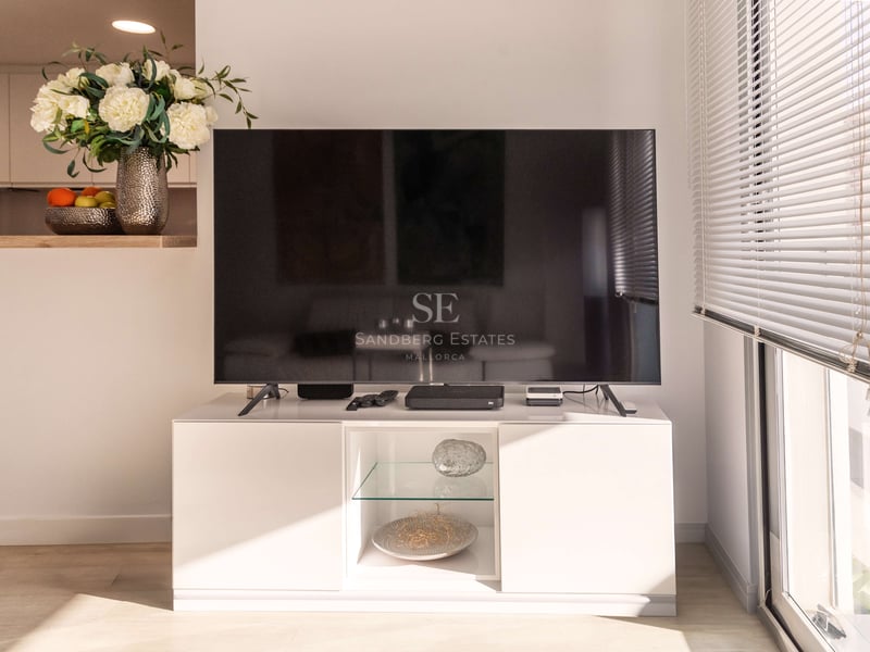 Flachbildfernseher auf weißem Lowboard neben sonnigem Fenster mit Jalousien und Blumendekoration.