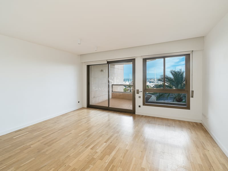 Tomt modernt rum med ljusa trägolv och stora fönster med utsikt över en balkong och en marina med palmer.