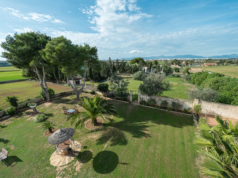 Vue surélevée d'une grande pelouse avec palmiers, murs en pierre et parasol en chaume avec les montagnes en arrière-plan.