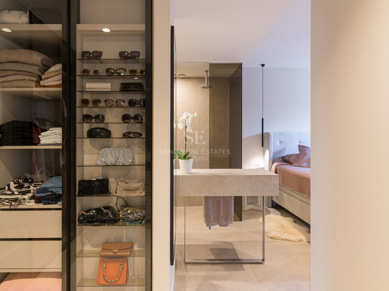 Modern walk-in closet with glass display shelves for accessories next to a stone vanity and master bedroom.