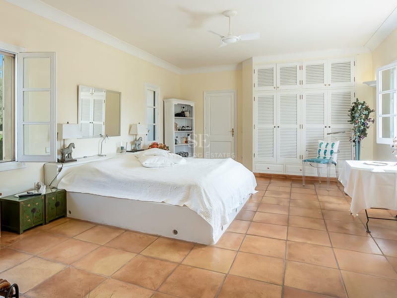 Chambre spacieuse avec sol en terre cuite, armoires à persiennes blanches et fenêtres donnant sur un jardin verdoyant.