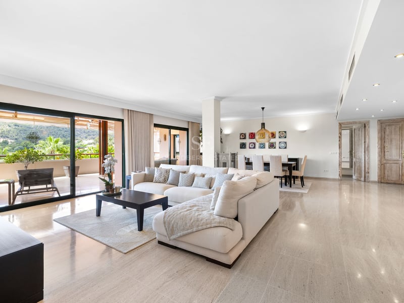 Salon spacieux avec canapé blanc en L, sol en marbre et grandes baies vitrées ouvrant sur une terrasse paysagée.