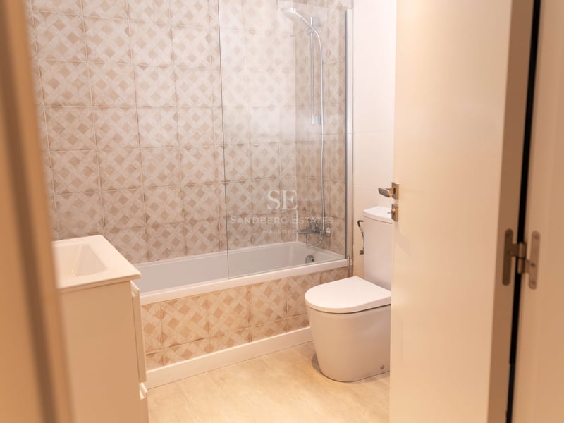 Badezimmer mit beige gemusterten Fliesen, Badewanne mit Glastrennwand, Toilette und Waschtisch.