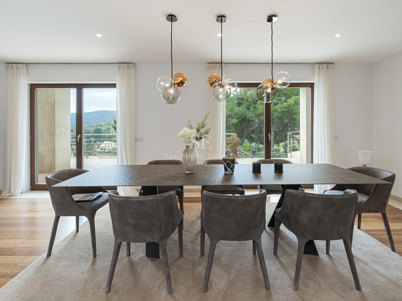Salle à manger moderne avec table texturée, chaises grises et suspensions en verre devant de grandes baies vitrées.
