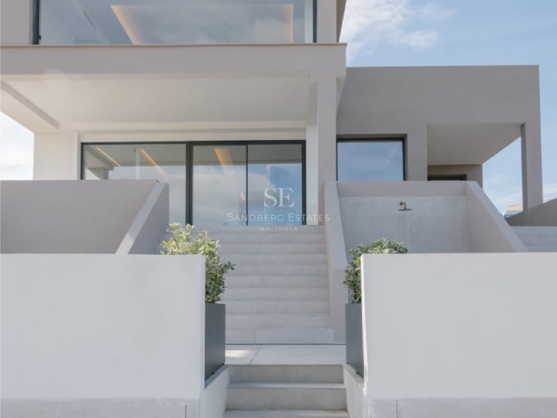 Vorderansicht einer modernen sandfarbenen Villa mit Steintreppen und großen Glasfenstern unter blauem Himmel.