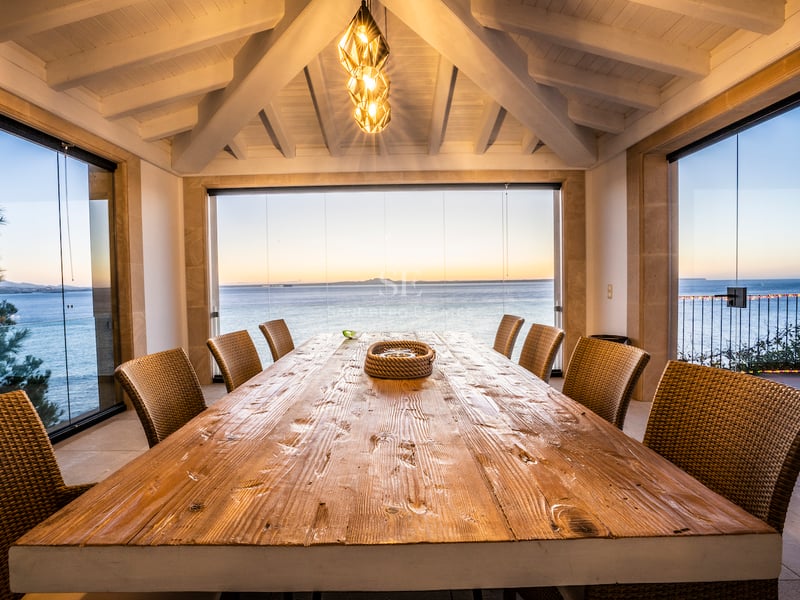 Großer Esstisch aus Holz auf einer verglasten Terrasse mit Panoramablick auf das Meer bei Sonnenuntergang.