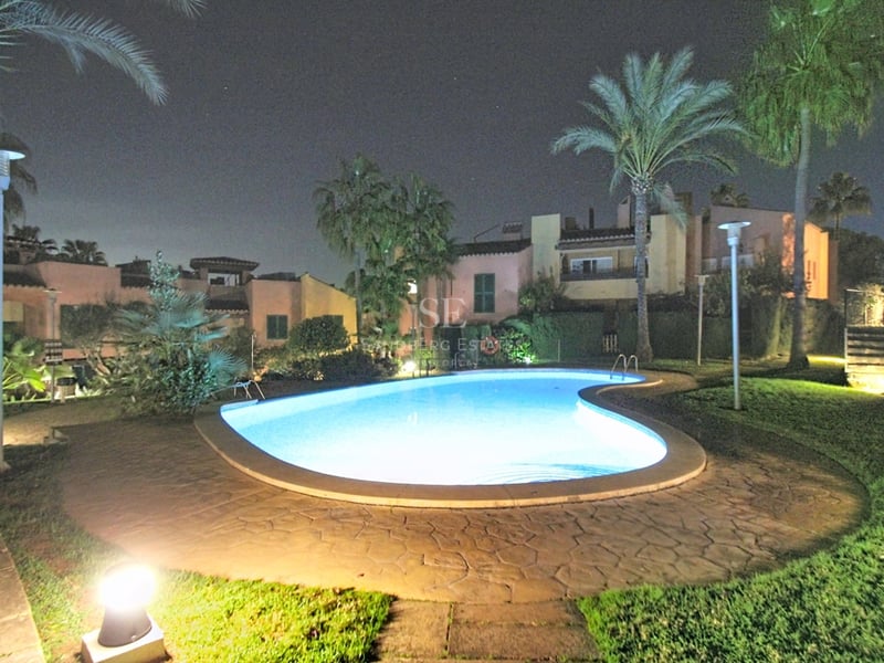 Njurformad pool upplyst på natten, omgiven av palmer, gräsmatta och medelhavsvillor under en mörk himmel.